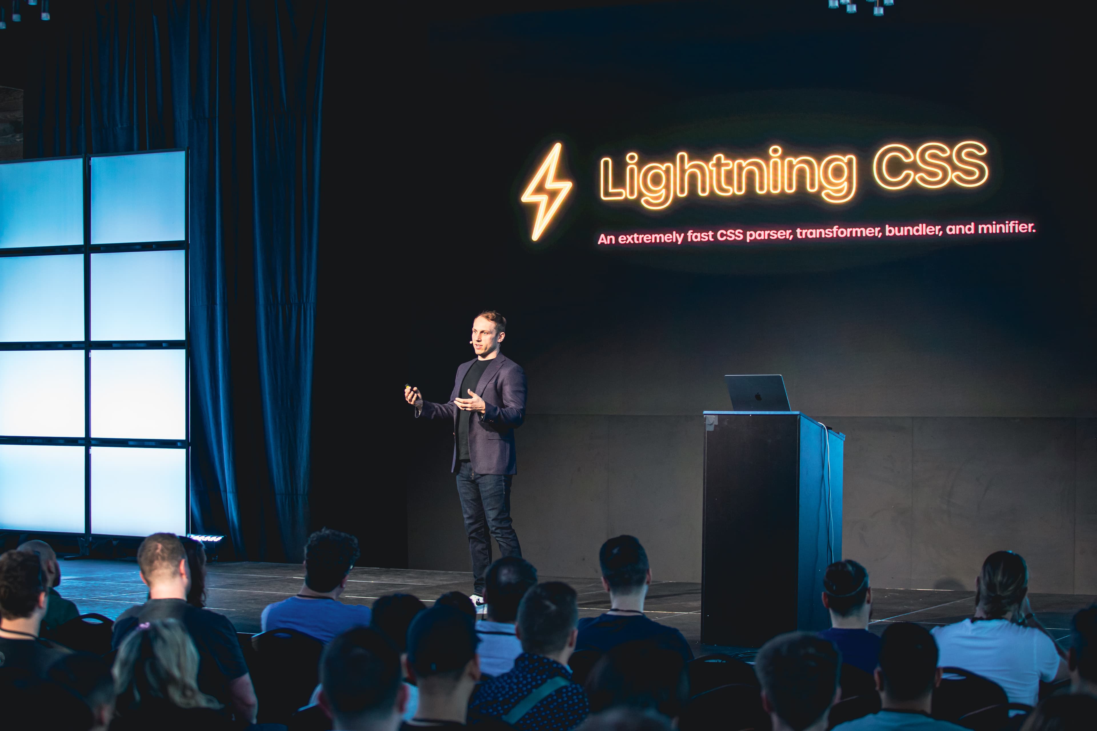 Adam Wathan 站在舞台上，面前有一张写着 Lightning CSS 的幻灯片